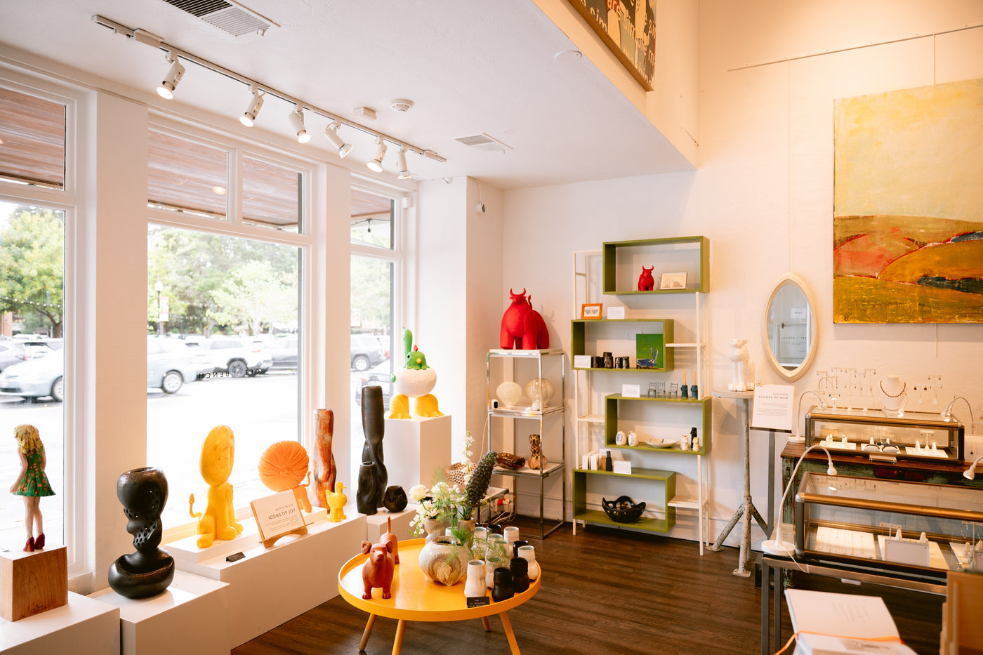 A bright, modern art gallery and design shop interior with large windows, white walls, and display shelves filled with sculptures, books, and decorative objects. A round yellow table sits in the foreground, and a framed abstract painting hangs on the right wall.