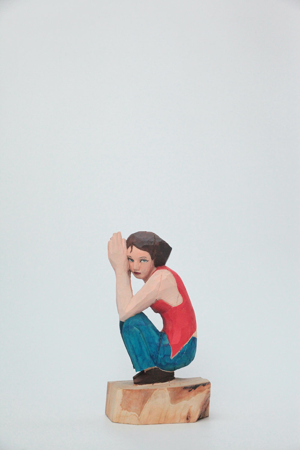 Artist Hideki Iinuma's wood sculpted female figure Jessica holds space in a contemplative pose, squatting, with her hands pressed together and gazing slightly downward.