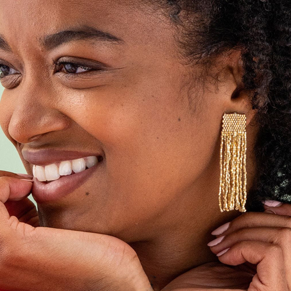 A close-up lifestyle shot of a woman wearing a long, gold beaded tassel or fringe earring. The earring features a woven rectangular top and cascading strands of small gold beads.