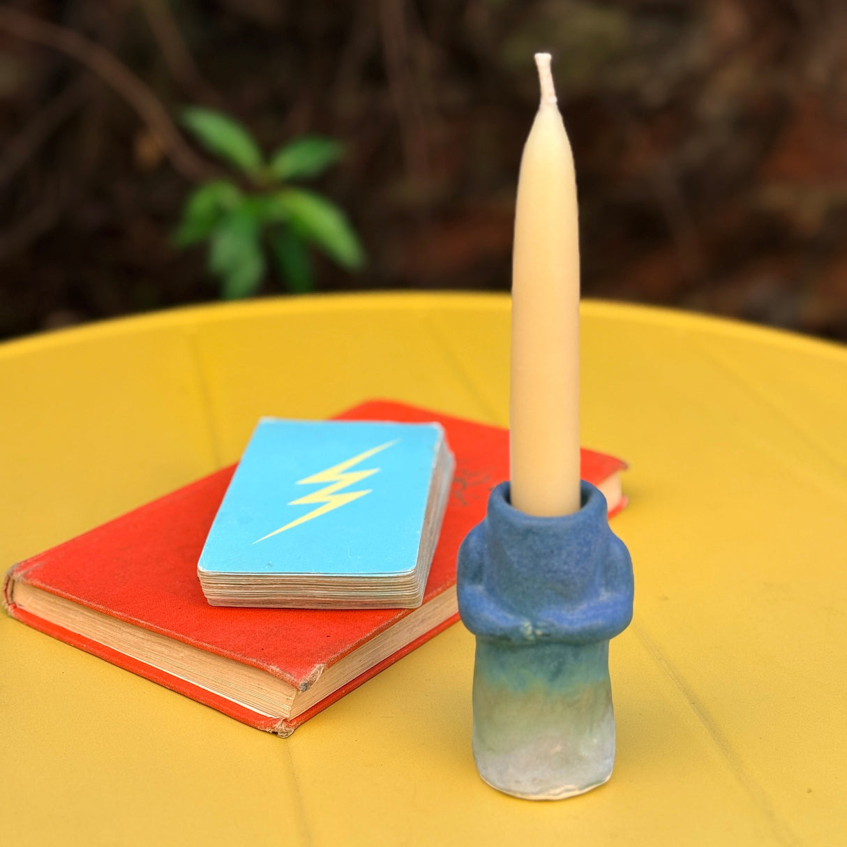 Kiki Plz Hug Candle in an ombre cobalt blue glaze with a candle, on a yellow surface with a deck of cards and a book in the background