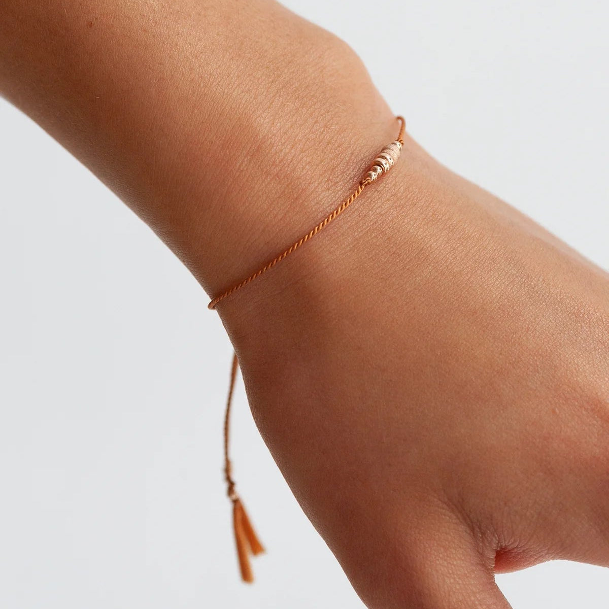 A delicate rose gold beaded and silk cord bracelet worn on a person's wrist, featuring a small tassel detail, shown against a plain light gray background.