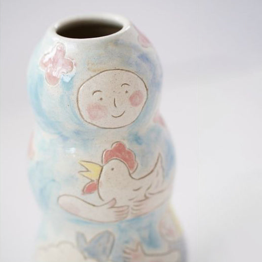 A tall flower vase is wheel thrown and decorated with characters in a scene: mom, child and chicken.