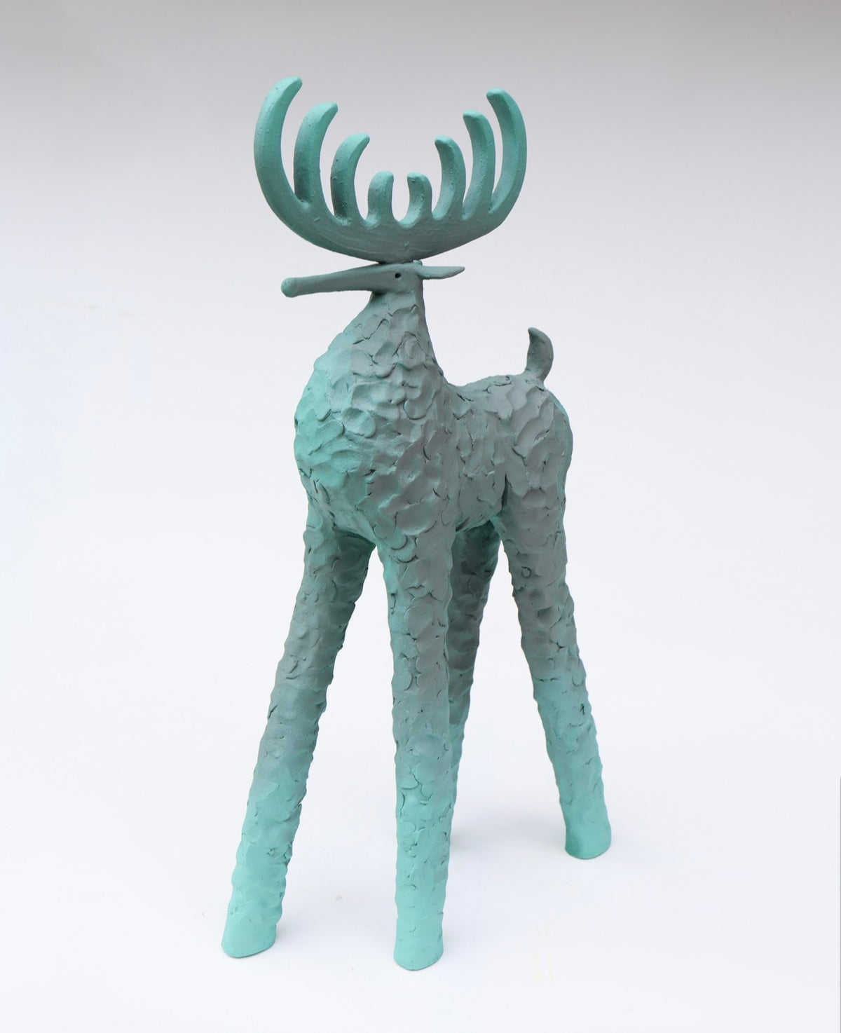 A stylized deer sculpture with textured, surfaces and large antlers, painted in a muted ombre teal-green color.