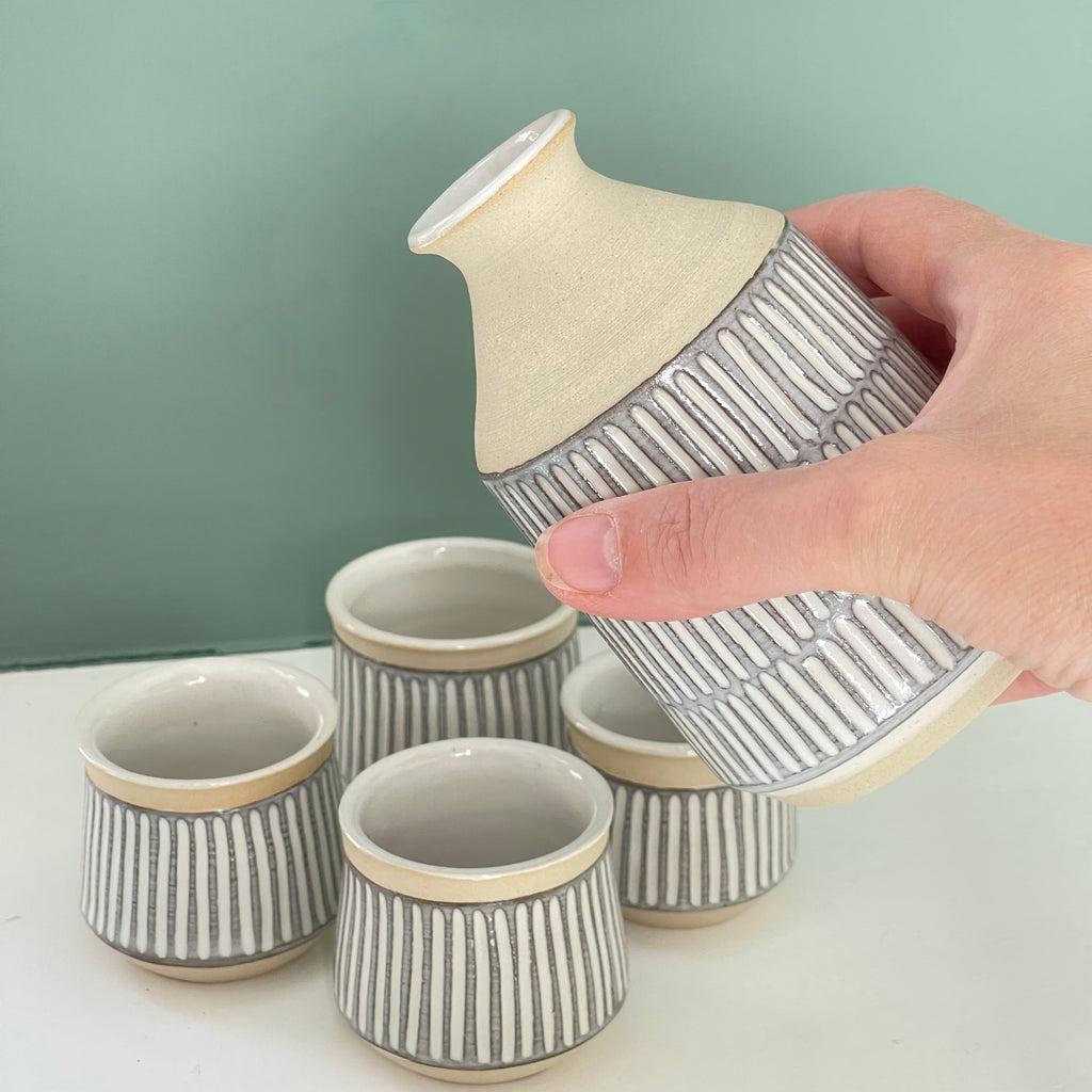 Handmade sake bottle, with different decorative techniques. Sgraffito technique, where lines are carved through a layer of dark colored slip, finished off with a white glaze for a light profile. Shown as a set with a bottle being poured and sake cups. 