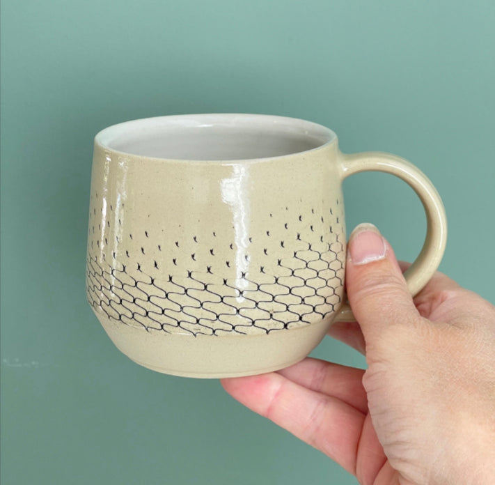 Get cozy with a warm cup, wheel thrown and decorated with a Scandinavian aesthetic. This substantial beauty of a mug comes in 14oz to hold your favorite beverage or soup.  This mug is imprinted with a netting relief, creating a gradient on the body—more defined at the bottom and disappearing higher up. The netting imprint is colored black and glazed in a transparent glaze, while the inside of the mug is glazed in white.