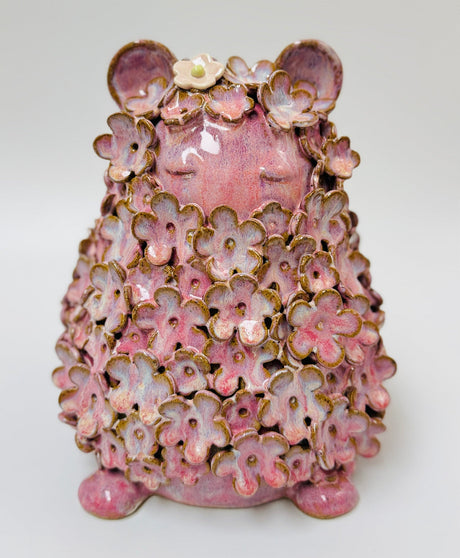 A pink ceramic figurine in a meditative sitting pose, covered in layered pink cherry blossom petals with gold-toned edges