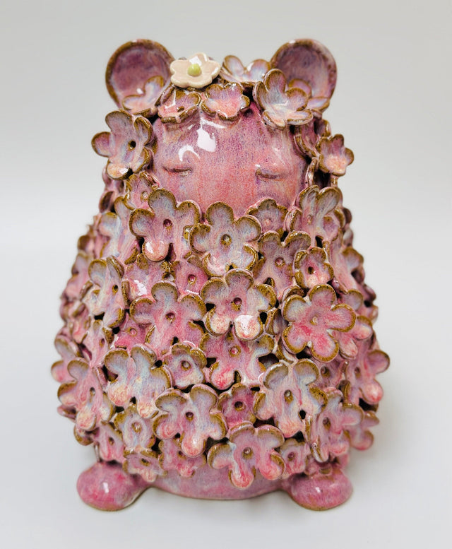 A pink ceramic figurine in a meditative sitting pose, covered in layered pink cherry blossom petals with gold-toned edges