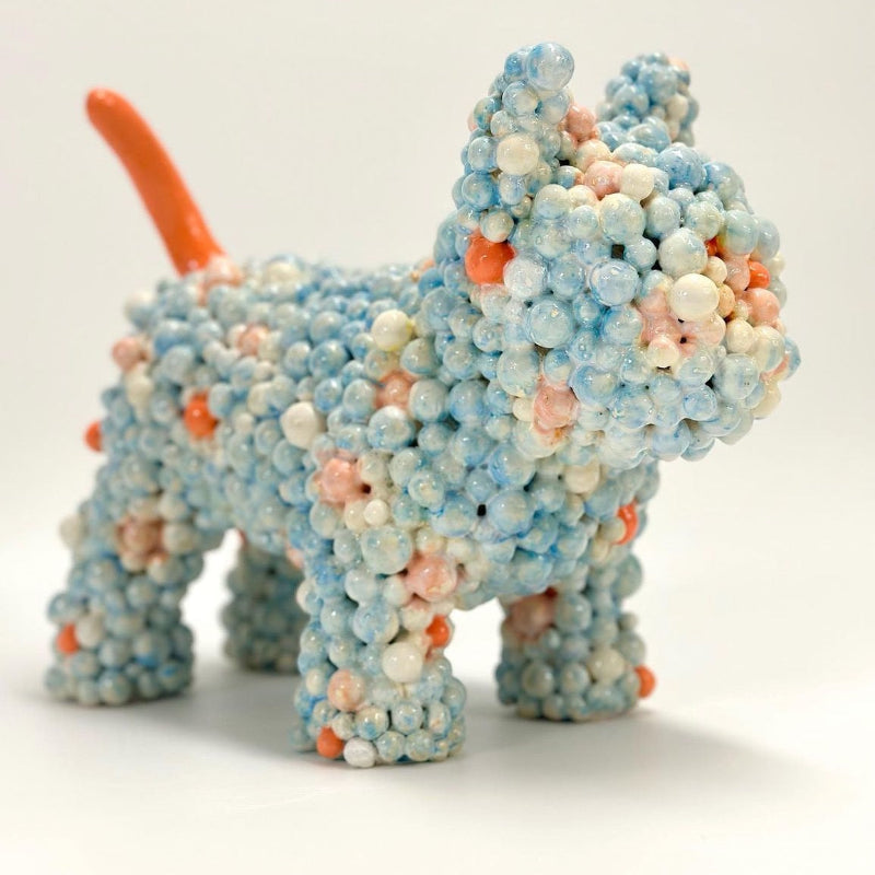 A ceramic dog sculpture made of small blue, white, and peach indivdually sculpted bubble gum balls, with a bright orange tail.