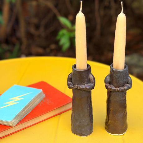 Two black hug candle holders with candles on a yellow surface with books and greenery in the background