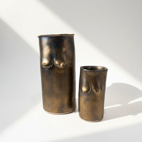 Two bronze-colored cylindrical vases and cup with sculpted breasts in ceramic on a white surface.