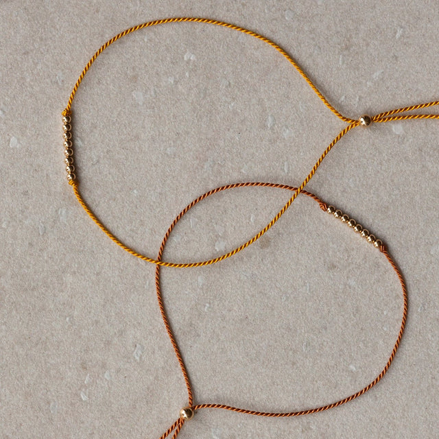 Close-up of thin braided silk cord friendship bracelets with small 14k gold filled bead accents and tassels