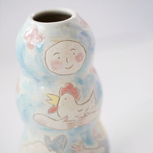 A tall flower vase is wheel thrown and decorated with characters in a scene: mom, child and chicken. 