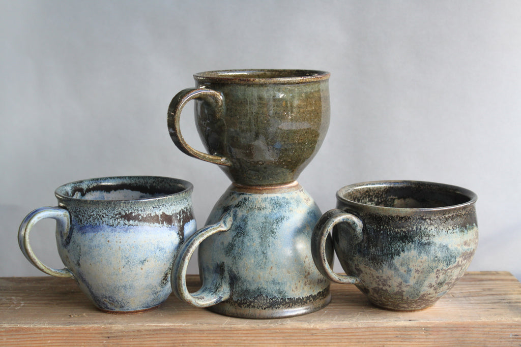 stoneware belly mugs. Rounded and tapered to feel nice in the hand, they have a one-finger handle. We love this variable blue glaze, like the ocean in your palm. Shown as a group with a dark green mug.